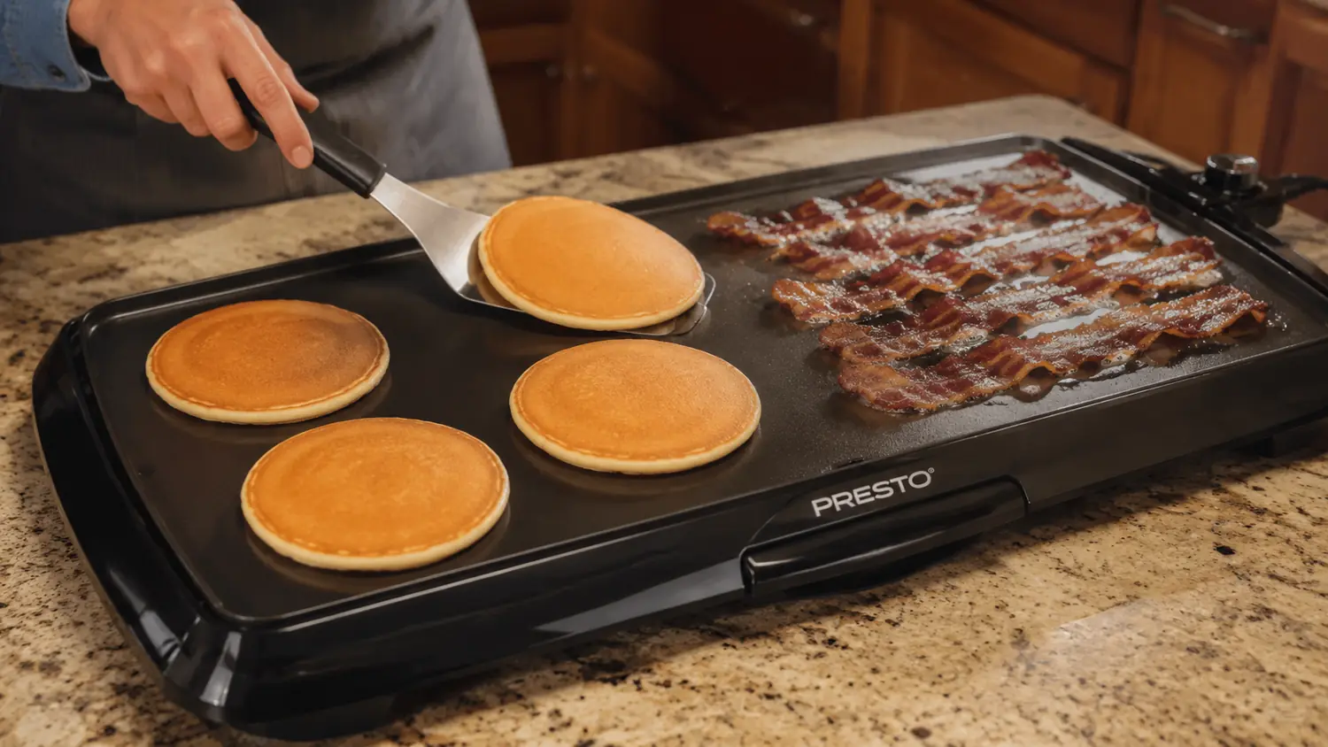 Hand cooking bacon, scrambled eggs, and pancakes at the same time on a large electric griddle
