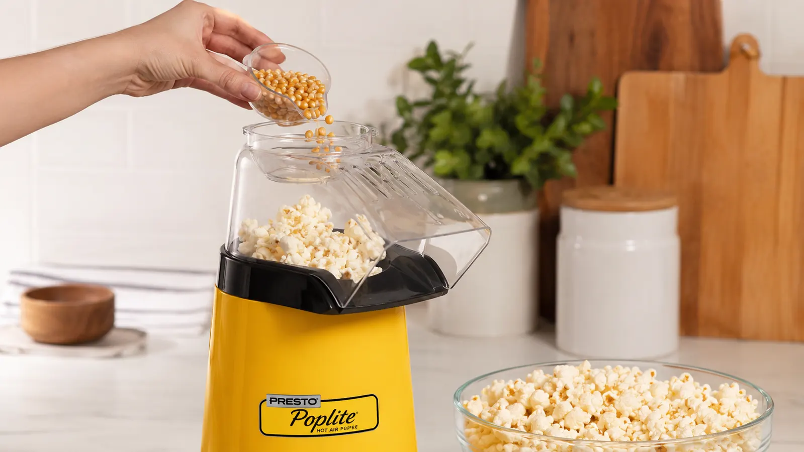 A hand pouring popcorn kernels into the measuring cup on top of the Presto Poplite hot air popcorn popper on a kitchen counter
