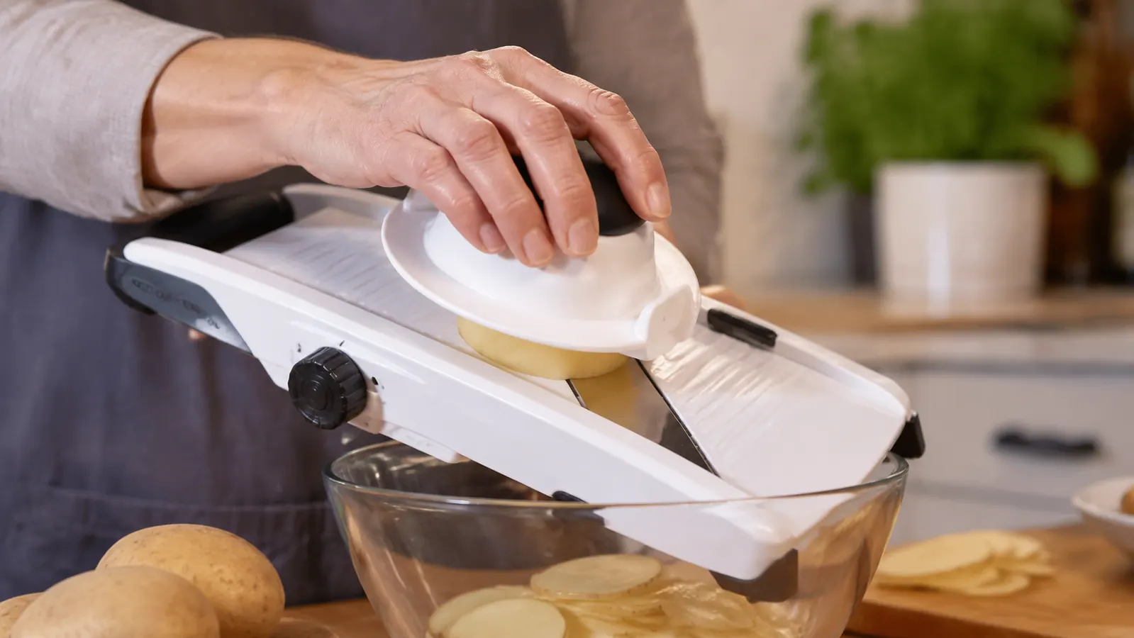 Hand using the plastic food holder to guide a potato safely across the mandoline blade over a glass bowl