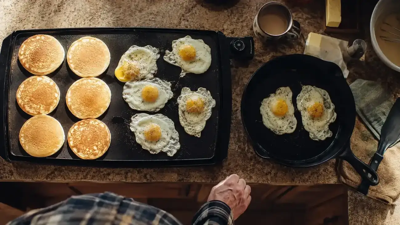 A black Presto 22-inch electric griddle sitting next to a well-seasoned cast iron skillet on a kitchen counter, both loaded with breakfast food